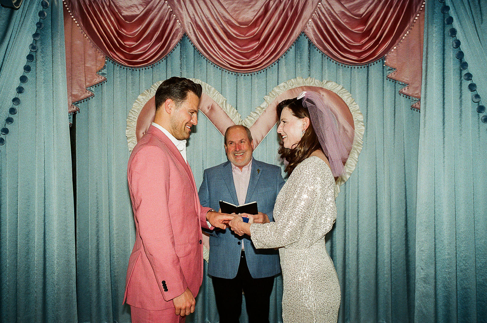 Couple exchanging rings during a Las Vegas chapel wedding ceremony, groom in a pink suit and bride in a sequined dress with veil, standing in front of a pastel heart-shaped backdrop with an officiant behind them