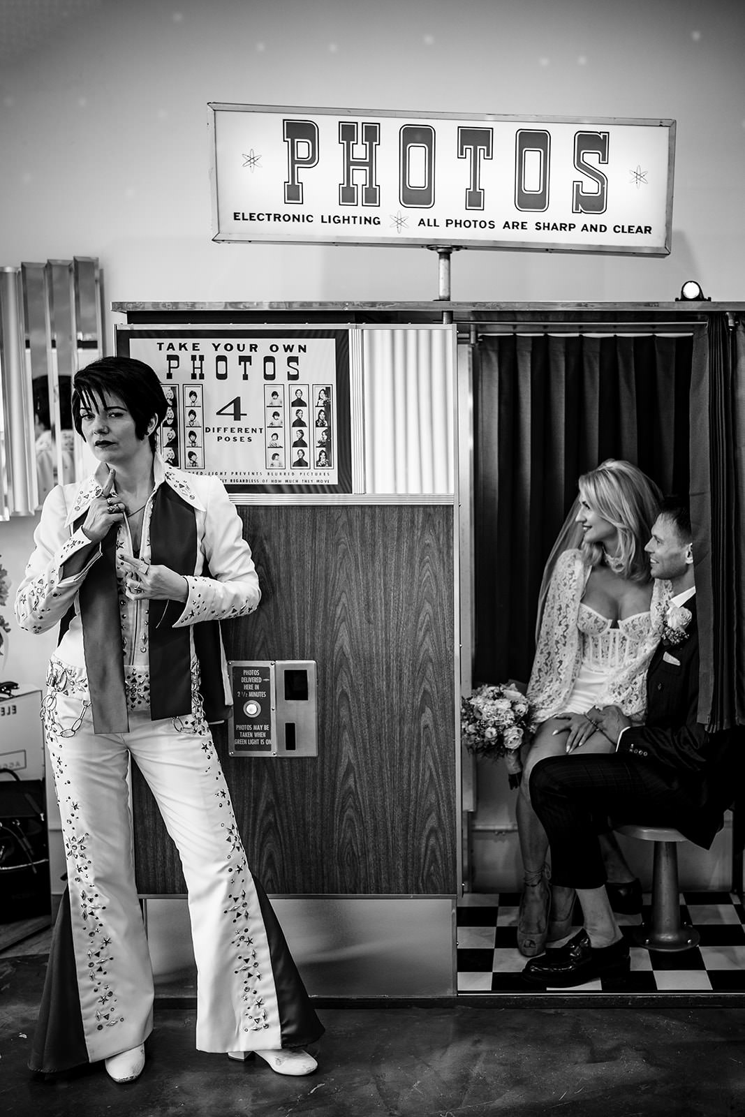 Black and white photo of an Elvis impersonator adjusting his outfit beside a vintage photo booth, while a bride and groom sit inside smiling at each other during a Las Vegas wedding