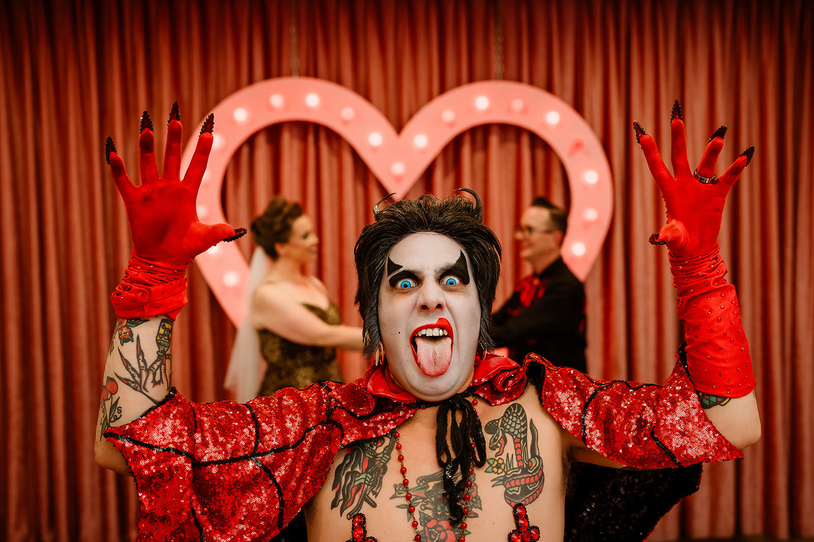 Hellvis officiant with dramatic makeup and red sequined cape posing in front of a lit heart backdrop while a couple exchanges vows in the background at Sure Thing Chapel in Las Vegas