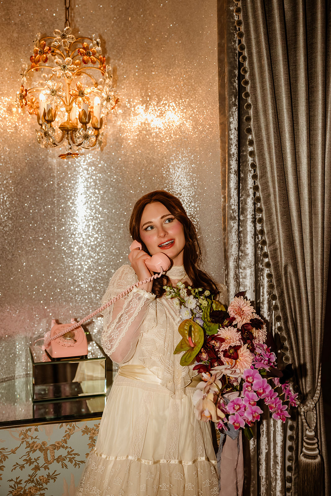 Bride in a lace wedding dress holding a pink vintage telephone and bouquet in a glam room with a chandelier and sparkling wall at Sure Thing Chapel in Las Vegas