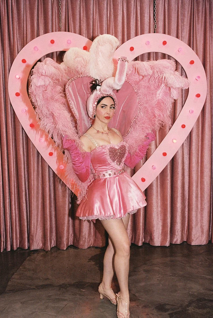 Pink showgirl performer posing in front of a lit heart-shaped backdrop with feathers and satin curtains at Sure Thing Chapel in Las Vegas