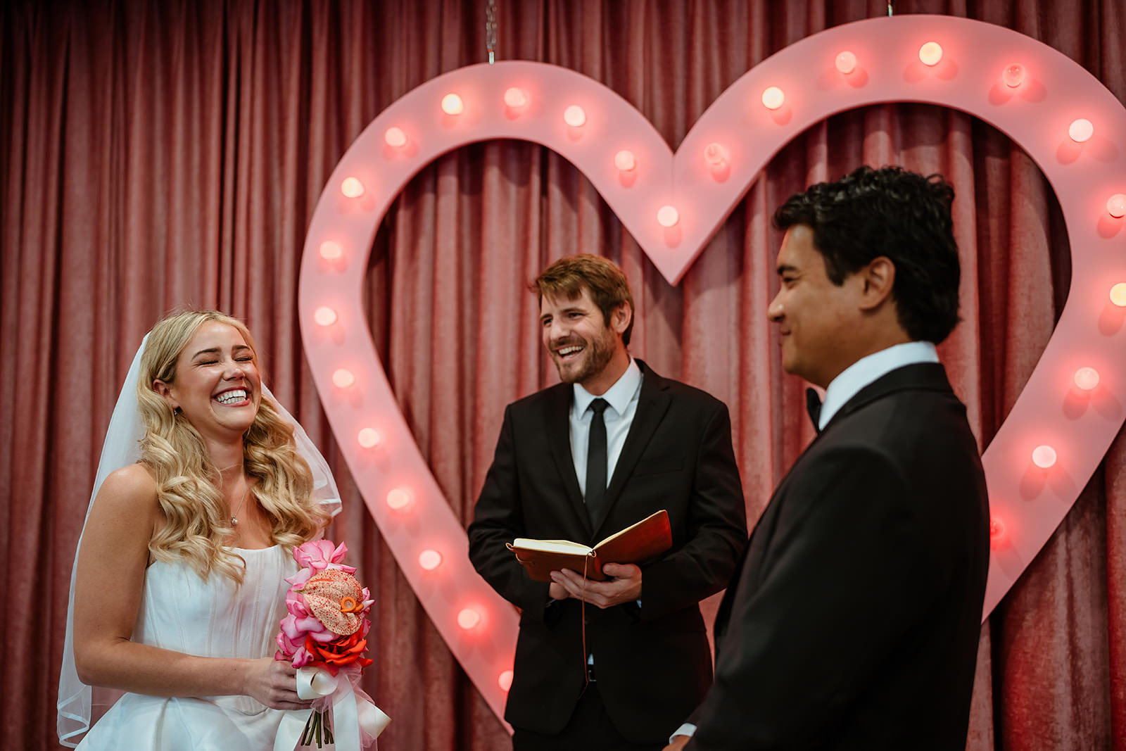 Bride and groom smiling during their wedding ceremony with an officiant in front of a lit heart-shaped backdrop at Sure Thing Chapel in Las Vegas