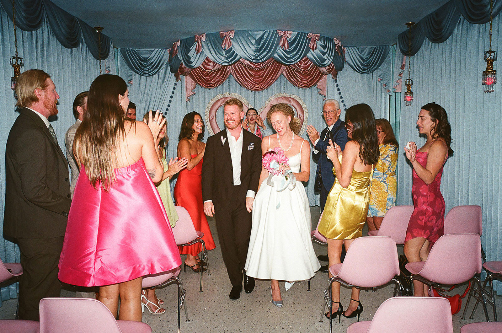 Newly married couple walking down the aisle as guests cheer and clap inside Sure Thing Chapel in Las Vegas, with pastel drapery and heart backdrop behind them